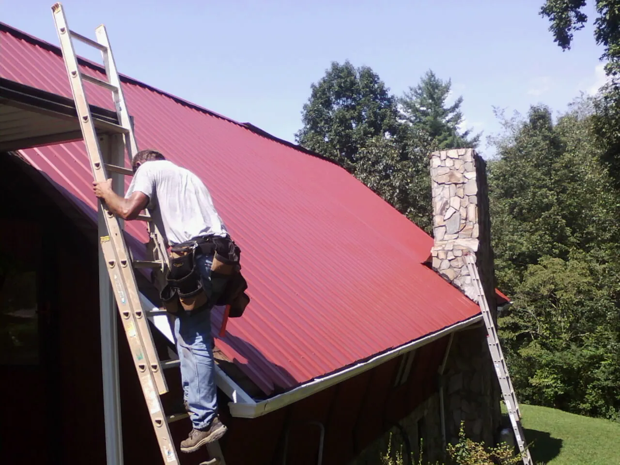 Roofer on a ladder beside a red metal roof and stone chimney — Soffit & Fascia Repair by Roofing Brothers in Donald