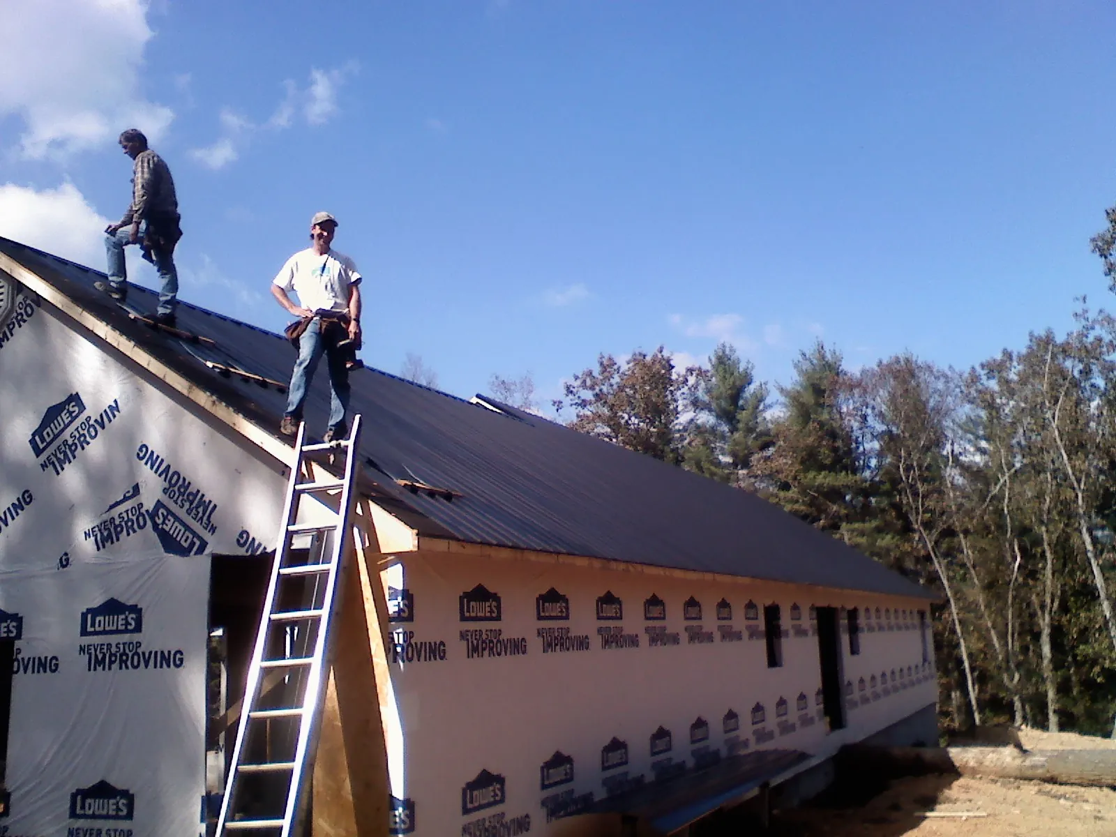 Two roofers standing on a long dark metal roof over a wrapped building in Donald